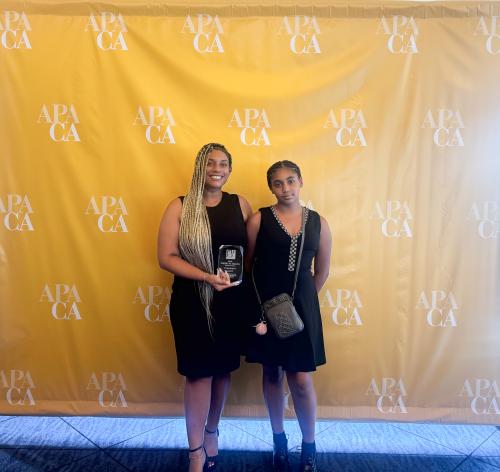 Two women in black dresses stand smiling in front of a yellow backdrop with a logo.